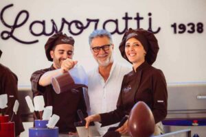 Fabio Fazio e due cioccolatieri, un uomo e una donna, uovo di Pasqua sul tavolo e sul retro scritta "Lavoratti"